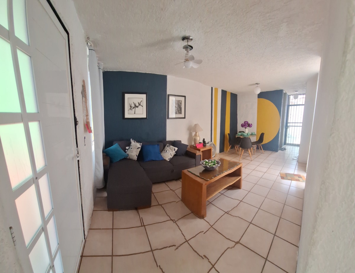 A welcoming living area features a sectional sofa adorned with decorative pillows, complemented by a wooden coffee table. A dining space is visible in the background, with a table and chairs. The walls are accented in shades of blue and yellow, enhancing the room's ambiance.