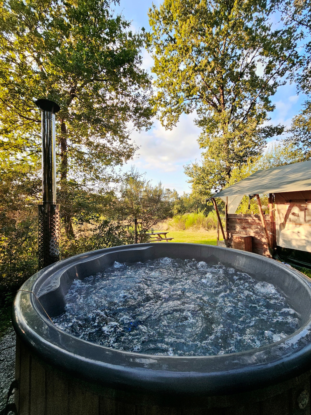 A wood-heated jacuzzi is filled with bubbling water, surrounded by lush greenery and trees. In the background, a safari tent is visible, set within a peaceful campsite. Natural light filters through the leaves, creating a serene outdoor environment.