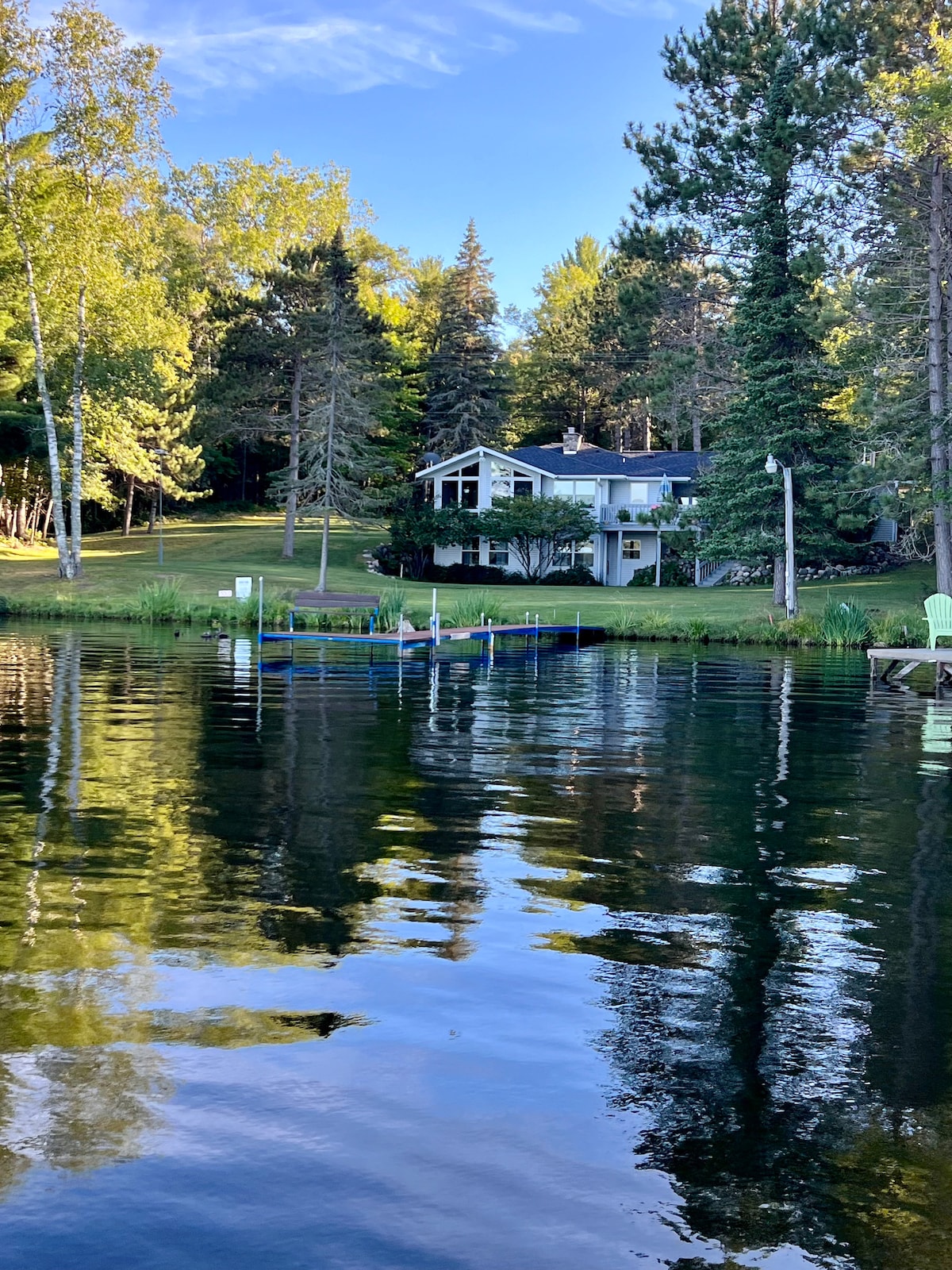 The lakeside view captures a charming house nestled among tall trees, reflecting in the tranquil waters. A small dock is visible, providing access to the lake, while lush green grass leads up to the home, enhancing the serene natural setting.