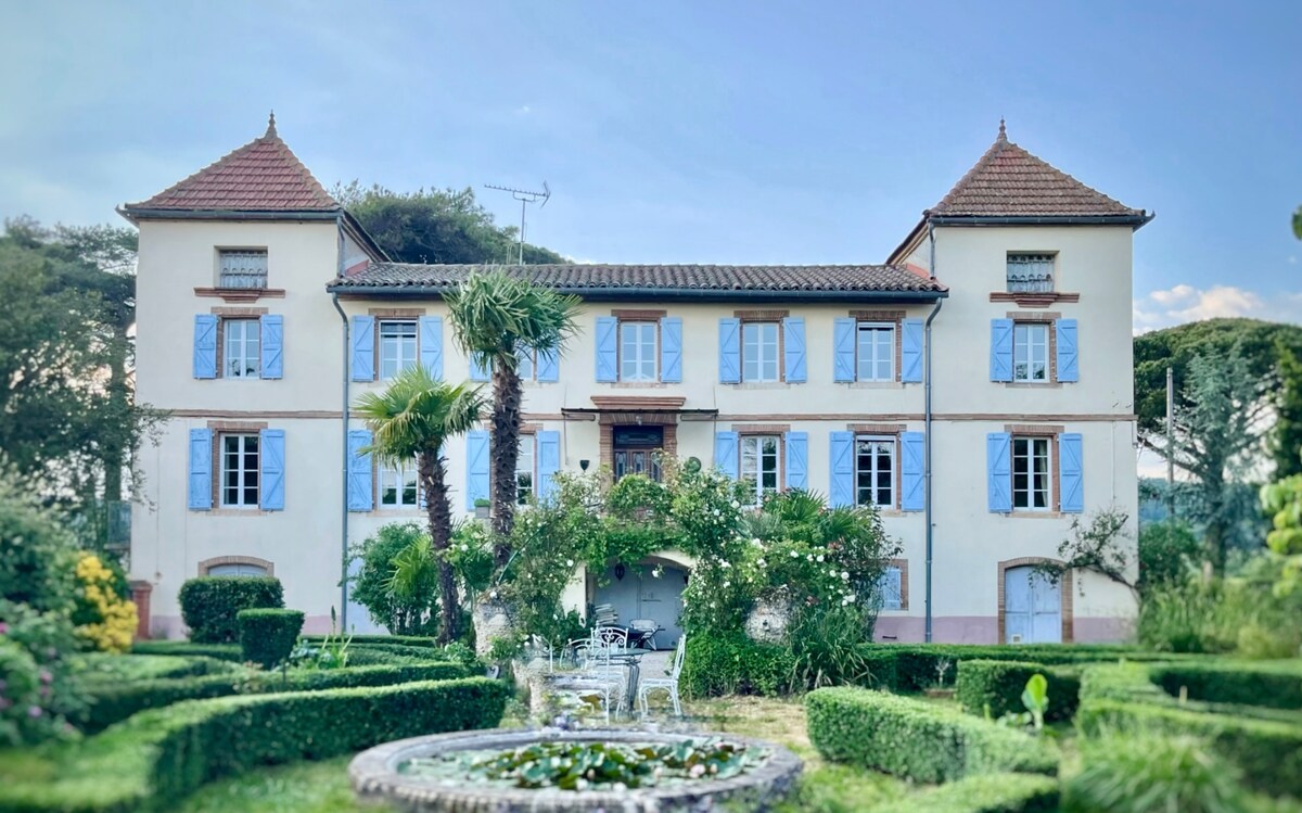 The charming façade of a historical stone building is framed by lush greenery and neatly trimmed hedges. Towering palm trees are visible in the garden, which features symmetrical flowerbeds and a decorative water fountain at the center.