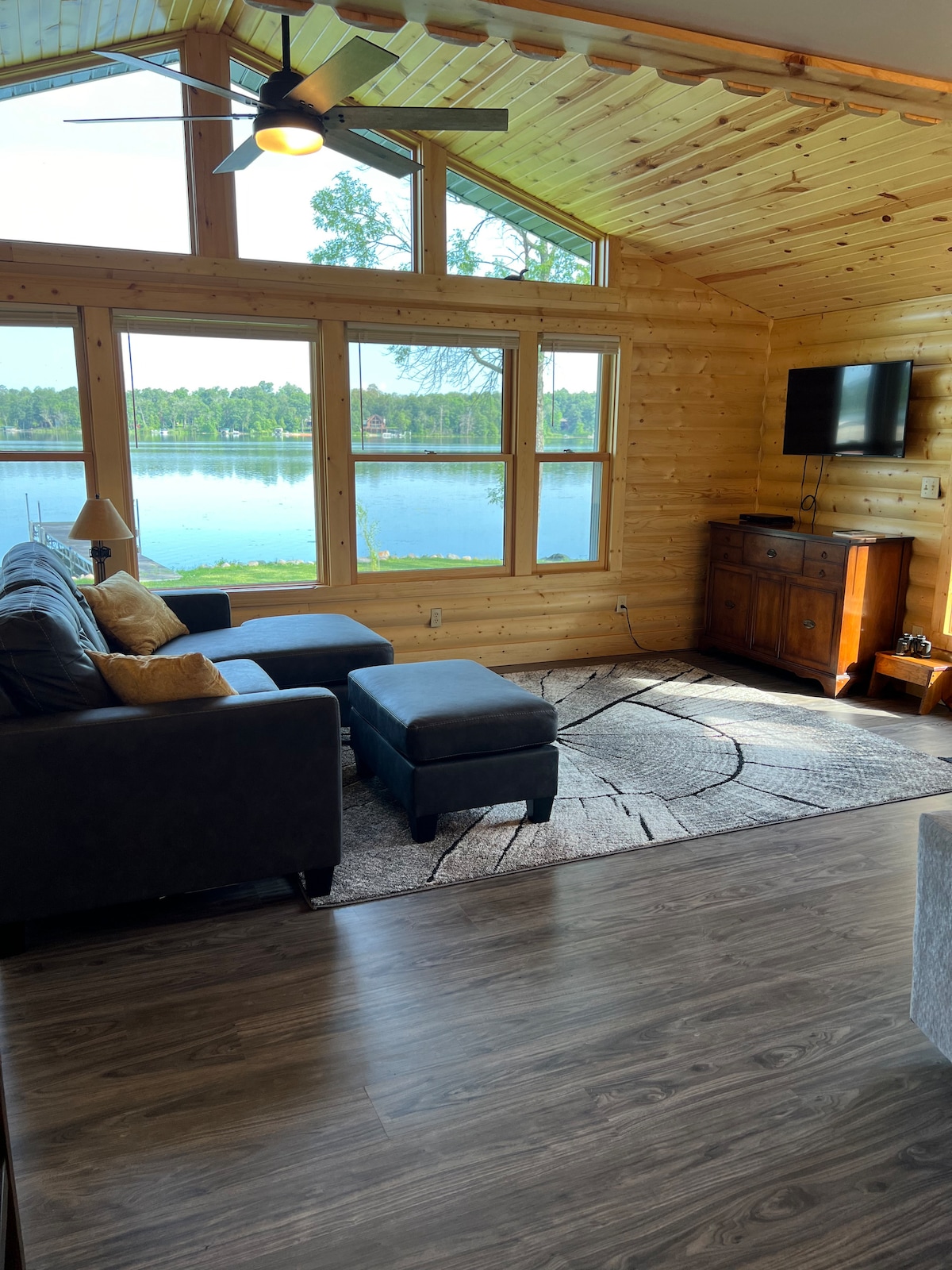 A spacious living area features wooden walls and ceiling, with large windows offering lake views. A comfortable dark sofa and an ottoman complement the space. Natural light fills the room, highlighting a media console and wall-mounted television.