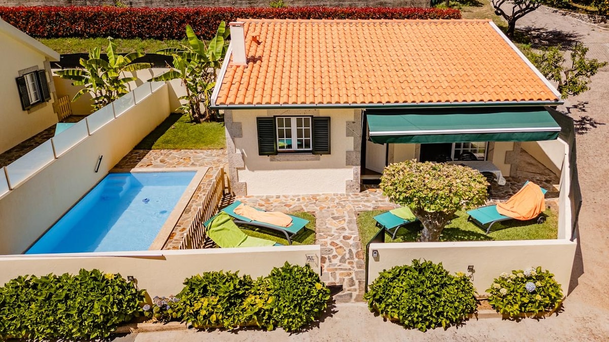 An aerial view showcases a charming house with a terracotta roof and green shutters. A private pool is visible alongside a stone patio featuring colorful lounge chairs. Lush greenery surrounds the property, creating a serene outdoor living space.