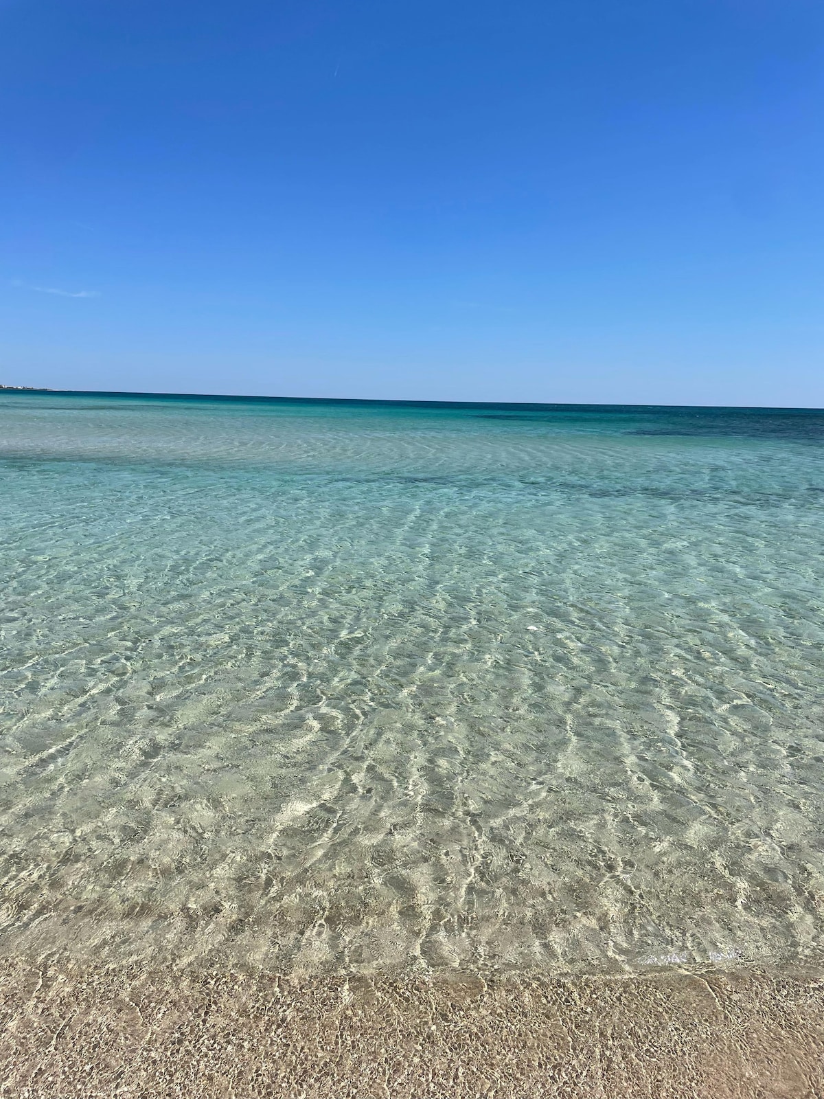 A serene beach scene is captured, showcasing clear, shallow water that reflects the blue sky above. The gentle waves are visible, creating ripples across the surface. Soft, golden sand borders the water's edge, leading to a tranquil coastal atmosphere.