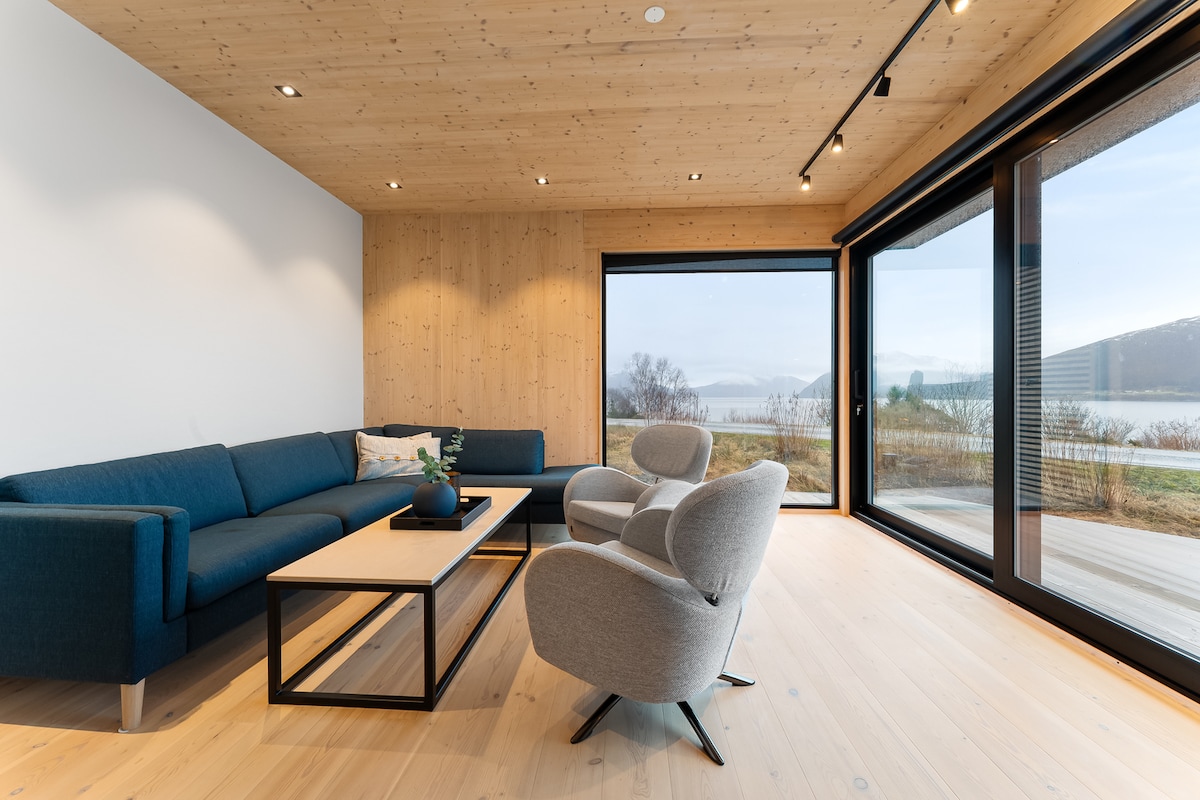 A bright living room features a cozy blue sectional sofa and light grey armchairs arranged around a modern coffee table. Large sliding glass doors provide an unobstructed view of the serene landscape outside, allowing natural light to fill the space.