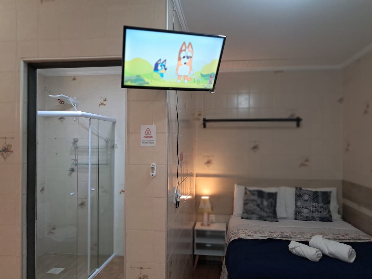 A compact room is presented, featuring a spacious bed with neatly arranged pillows and a cozy blanket. A modern television is mounted on the wall, showing animated content. A glass shower enclosure is visible in the bathroom area, with a soft light illuminating the space.