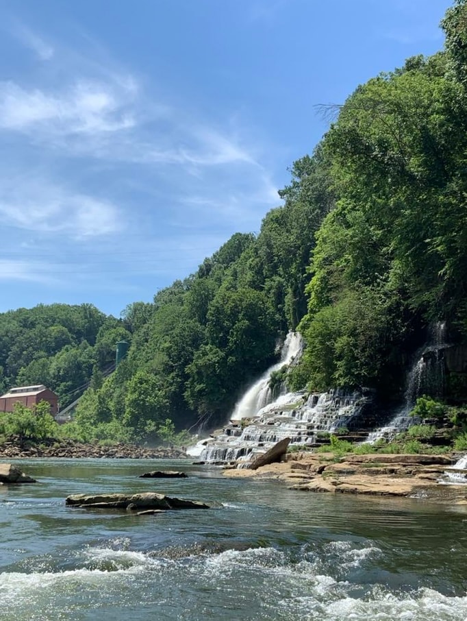 A serene view of a cascading waterfall is framed by lush green trees and rocky riverbanks. The water flows gracefully over layered rocks, creating a tranquil scene. In the background, a building is nestled among the trees, completing the natural landscape.
