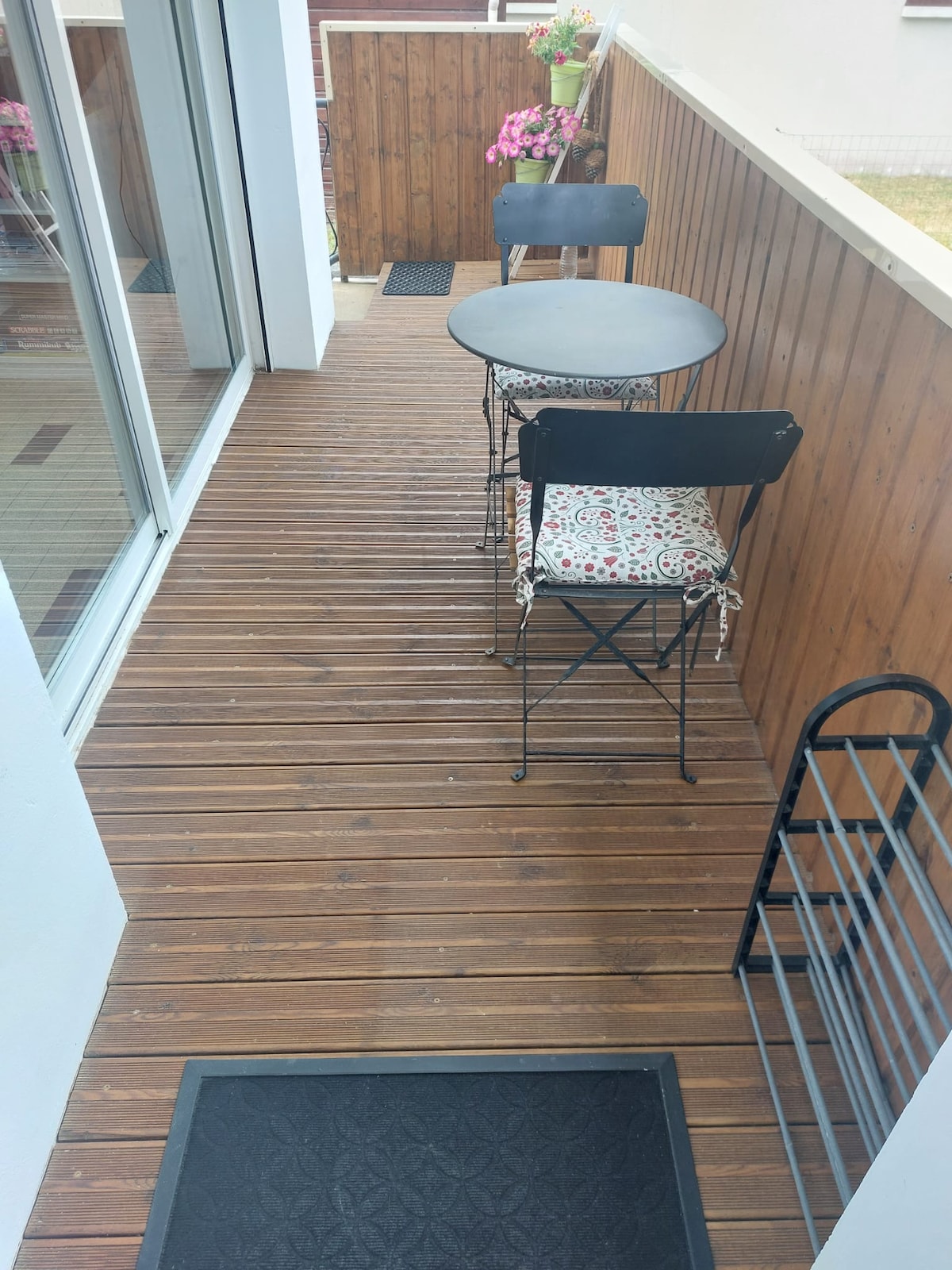 A small terrace area features a round table accompanied by two black folding chairs with decorative cushions. Wooden paneling decorates the walls, while potted flowers add a touch of color. A small rack is positioned nearby for convenience.