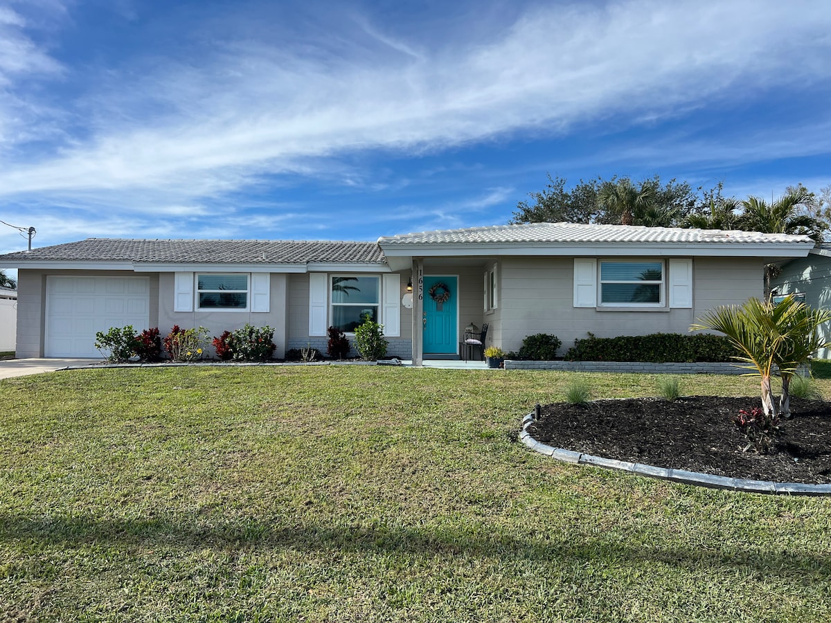 The exterior of a one-story lakefront home is showcased, featuring a well-maintained lawn with manicured shrubs and flowers. A bright turquoise front door stands out, complemented by a stone pathway and a small palm tree near the driveway. The sky is partly cloudy, adding a calm ambiance.