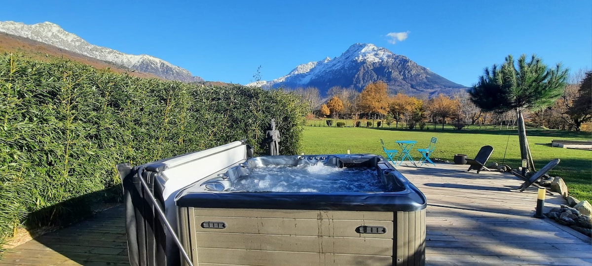 A private jacuzzi is situated on a wooden deck, with snow-capped mountains visible in the background. Lush greenery surrounds the area, and outdoor seating is arranged nearby, offering a serene setting for relaxation amidst nature.