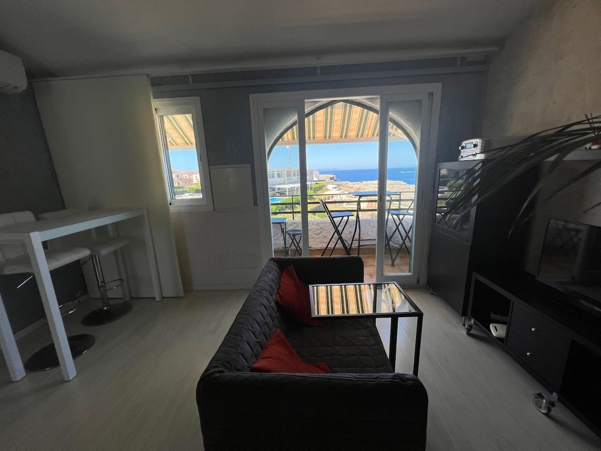 A comfortable living area features a glass-topped table and a dark sofa adorned with red cushions. Large glass doors open to a terrace, offering views of the sea. The space is complemented by bright natural light and a modern, minimalist design.