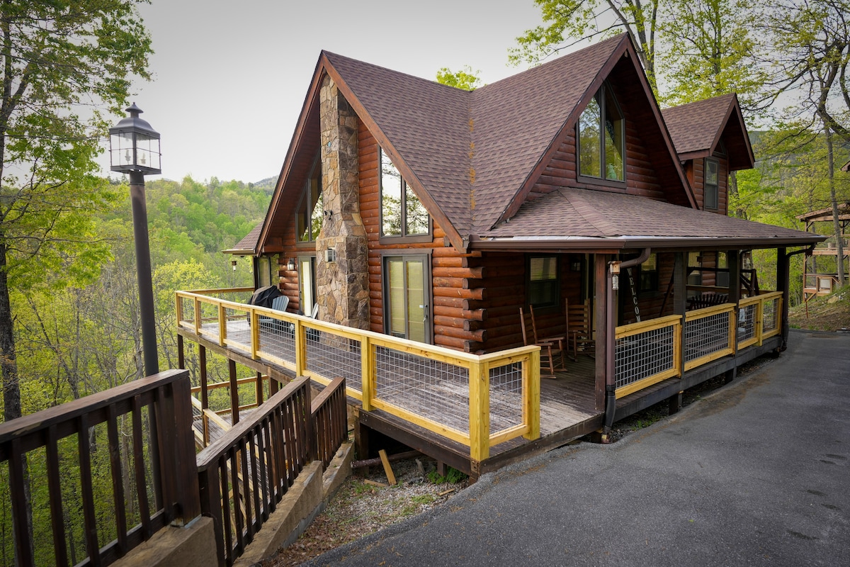 The three-level A-frame cabin features a rustic wooden exterior with a stone accent wall. A spacious wraparound deck is visible, surrounded by lush greenery. Large windows invite natural light, while outdoor seating provides a relaxing space to enjoy the surrounding landscape.