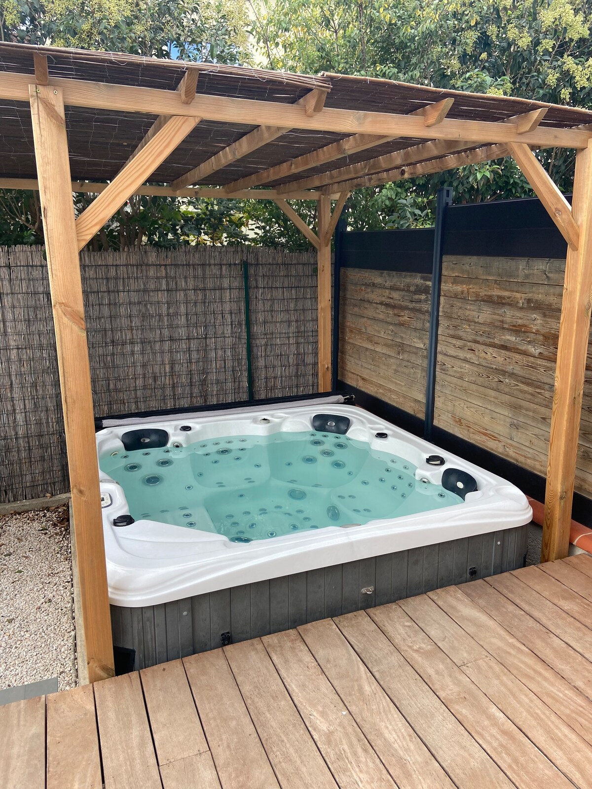 A private jacuzzi is situated beneath a wooden pergola, surrounded by a wooden deck. The spa features multiple seating areas with jets, providing a relaxing retreat. Lush greenery and a bamboo screen create a sense of seclusion and tranquility in the outdoor space.