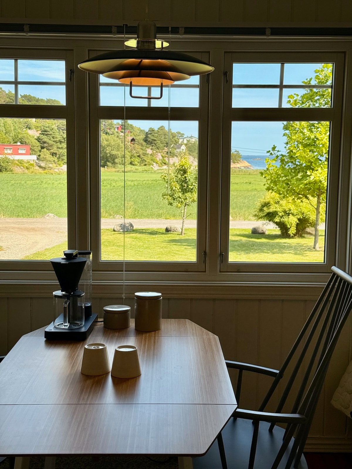 A dining table is positioned near a window, allowing views of the surrounding landscape and sea. Several cups and a coffee maker are arranged on the table. Soft light filters through the window, enhancing the calm ambiance of the space.