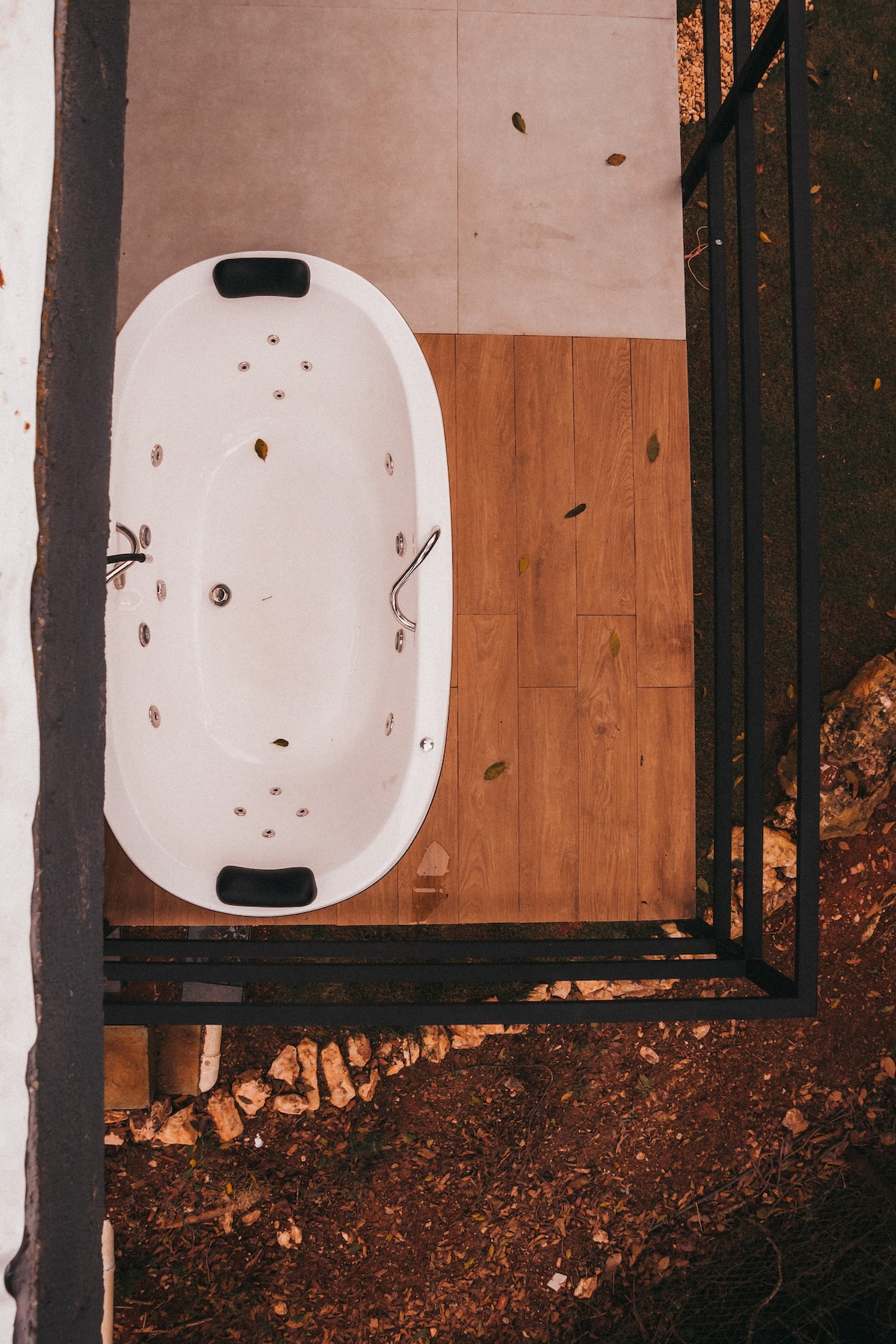 An outdoor tub is positioned on a deck surrounded by natural elements. The tub has a sleek white design with modern fixtures. The flooring combines light-colored tiles and wooden planks, while fallen leaves indicate a connection to the surrounding landscape.