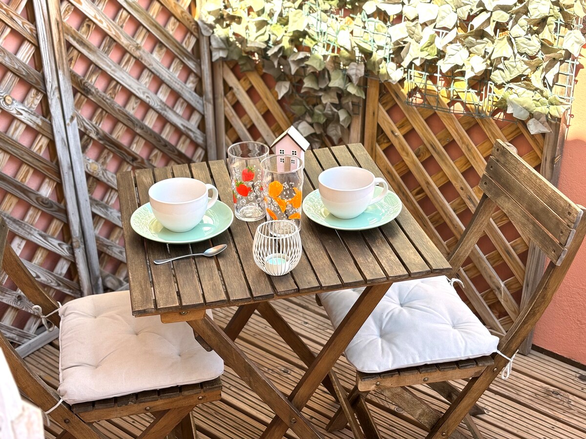 A wooden outdoor table is set for two, featuring pale green plates and matching bowls. Two cushioned chairs complement the table. The scene is adorned with decorative items including a glass, a beaded container, and a small colored centerpiece, surrounded by a trellis covered in foliage.