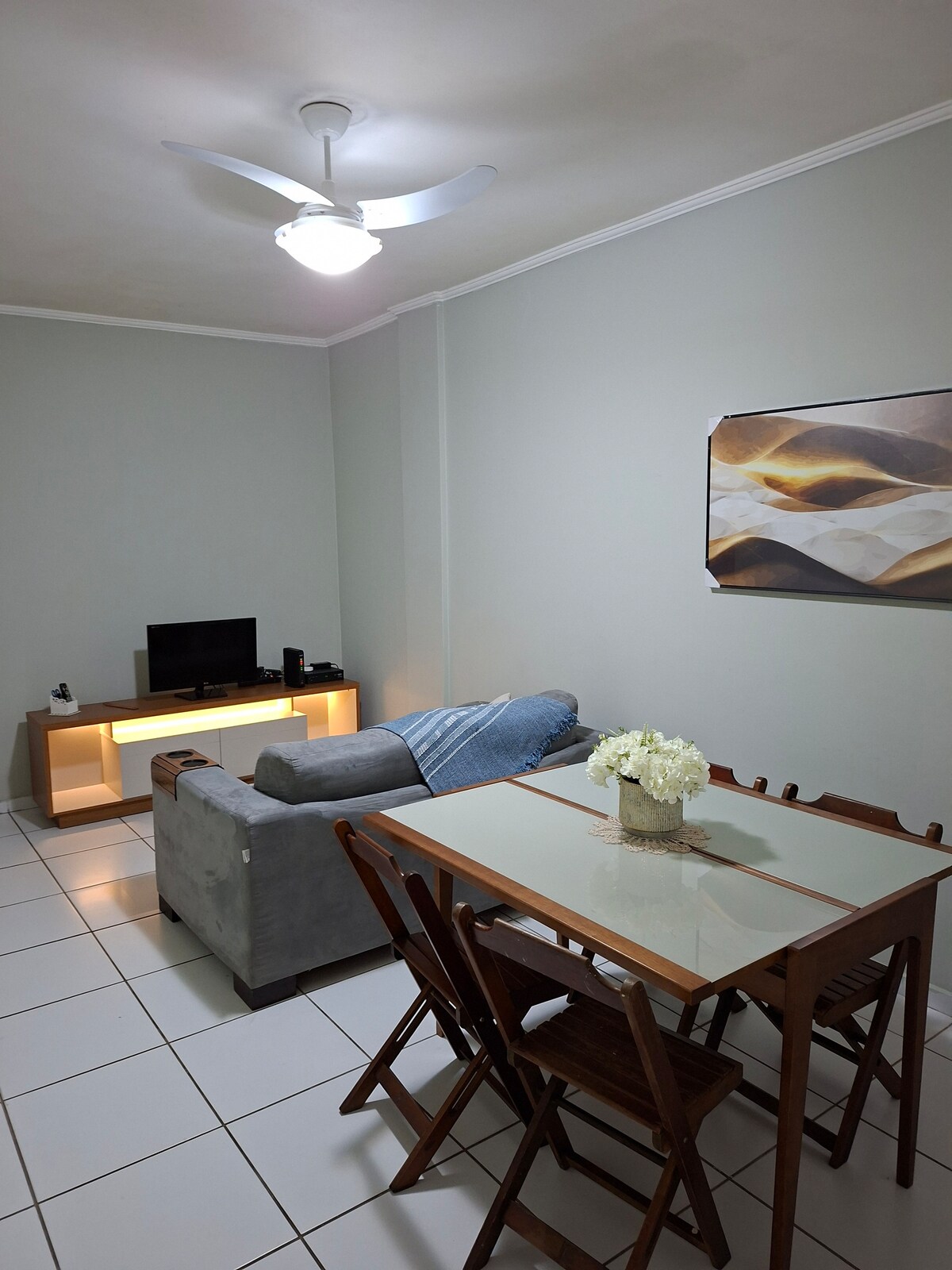 A cozy living area features a gray sofa, complemented by a light wood TV stand. A dining table with wooden chairs sits nearby, while a vase of white flowers adds a subtle touch. Soft lighting from the ceiling fan illuminates the space.