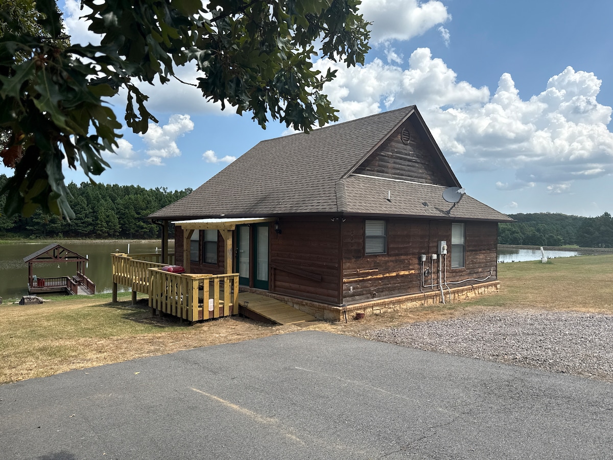 A log cabin-style home is depicted adjacent to a calm lake, featuring a wooden deck with seating. The exterior showcases natural wood tones, with a gravel area in the foreground. Lush greenery surrounds the peaceful setting, creating a connection with nature.