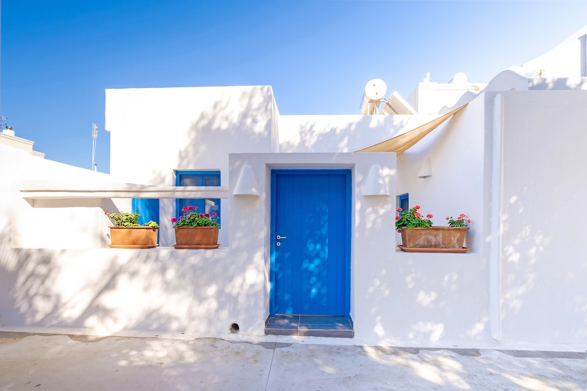 A charming entrance to a traditional Cycladic house is framed by white walls and a vibrant blue door. Flower boxes with colorful blooms are positioned beneath windows, enhancing the cheerful ambiance. Soft shadows from nearby trees add a serene touch to the inviting exterior.
