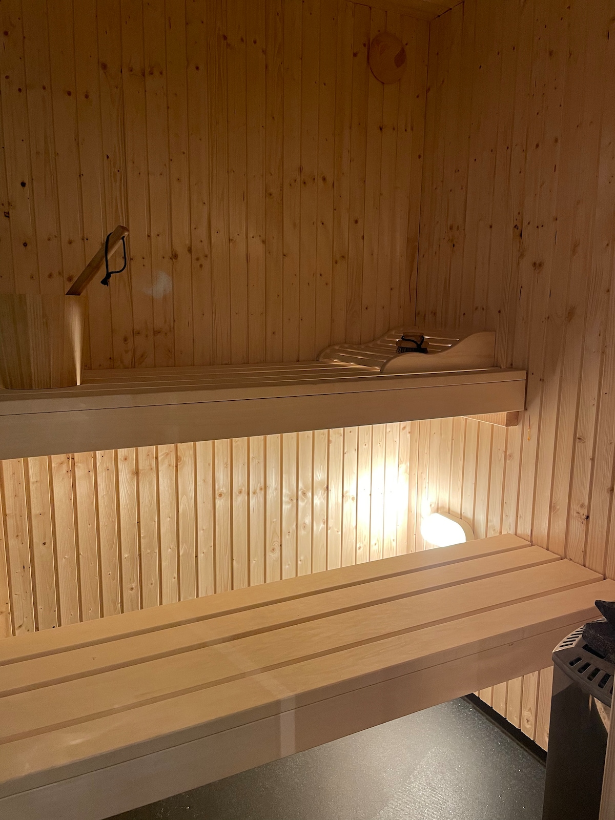 The interior of a wooden sauna is showcased, with two tiered benches made of light-colored wood. Soft lighting accentuates the warm tone of the walls, enhancing the soothing environment. A small window and a corner stove are also visible.