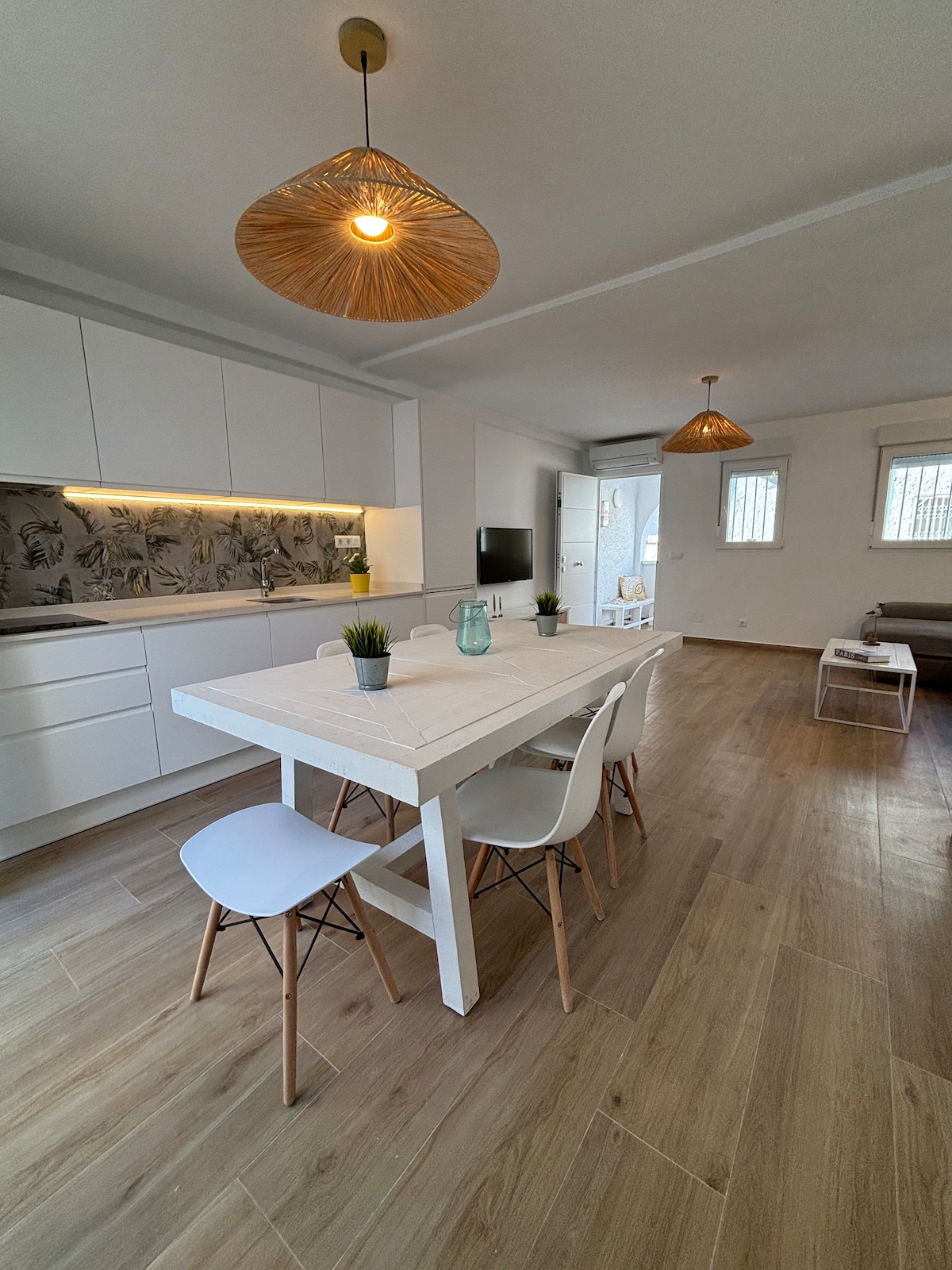 A bright kitchen space features a wooden dining table surrounded by four minimalist chairs. Overhead, two decorative light fixtures provide soft illumination. A modern kitchen with white cabinetry is visible, complemented by a patterned backsplash. A large window invites natural light into the room.