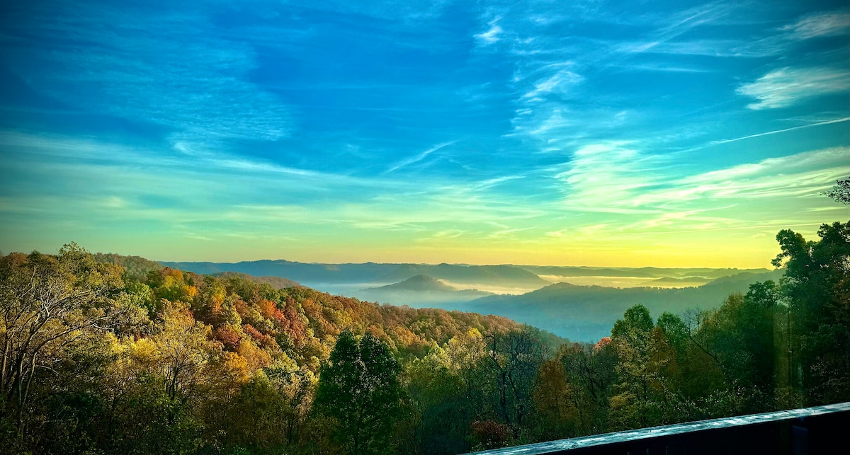 A panoramic view showcases rolling hills adorned with vibrant autumn foliage, blending shades of orange, yellow, and green. A hazy mist rises over valleys below, under a clear blue sky illuminated by the soft glow of dawn.