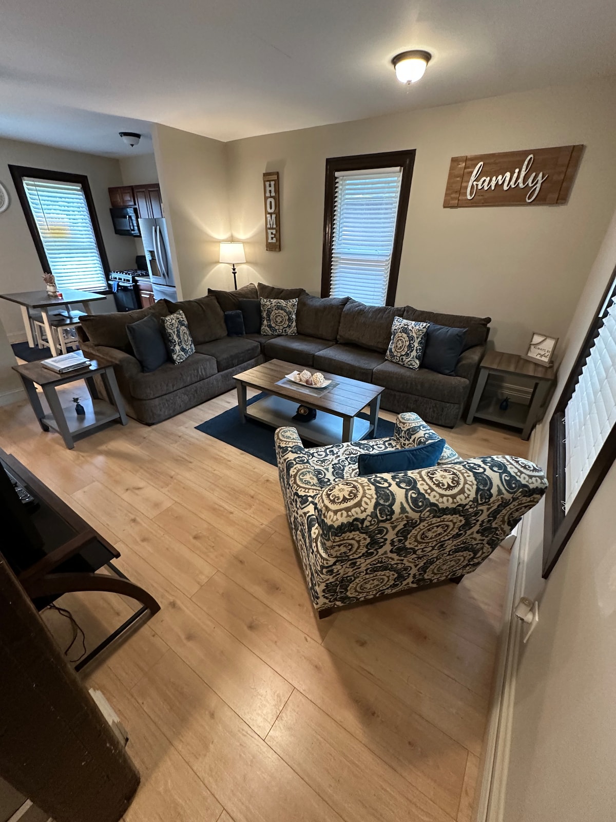 A comfortable living space features a sectional sofa adorned with patterned cushions, accompanied by a matching armchair. A coffee table sits centrally, while light streams in through the window, highlighting the warm wood flooring. Wall decor includes a 'family' wooden sign.