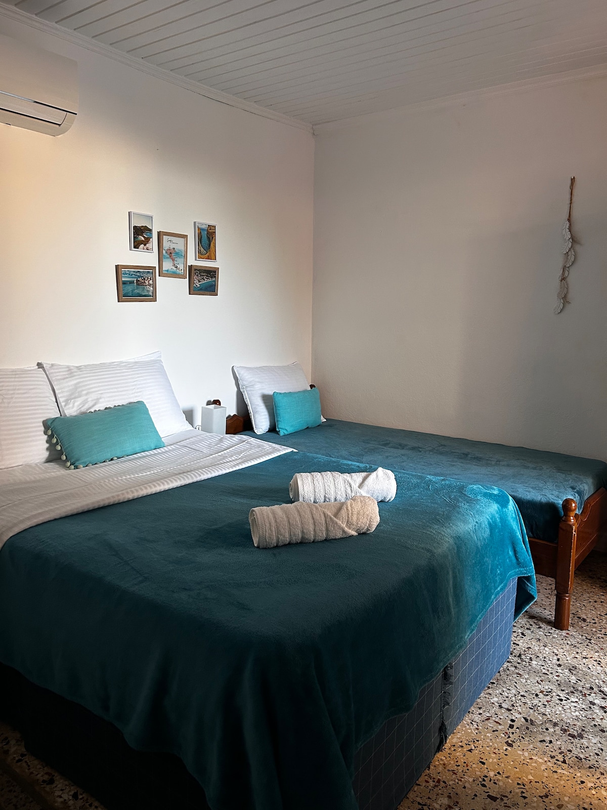 Two spacious beds are arranged with soft, textured blankets in a calming shade of blue. Freshly rolled towels are placed at the foot of each bed. Wall decor features a collection of framed photos, enhancing the ambiance. Natural light filters through the room.