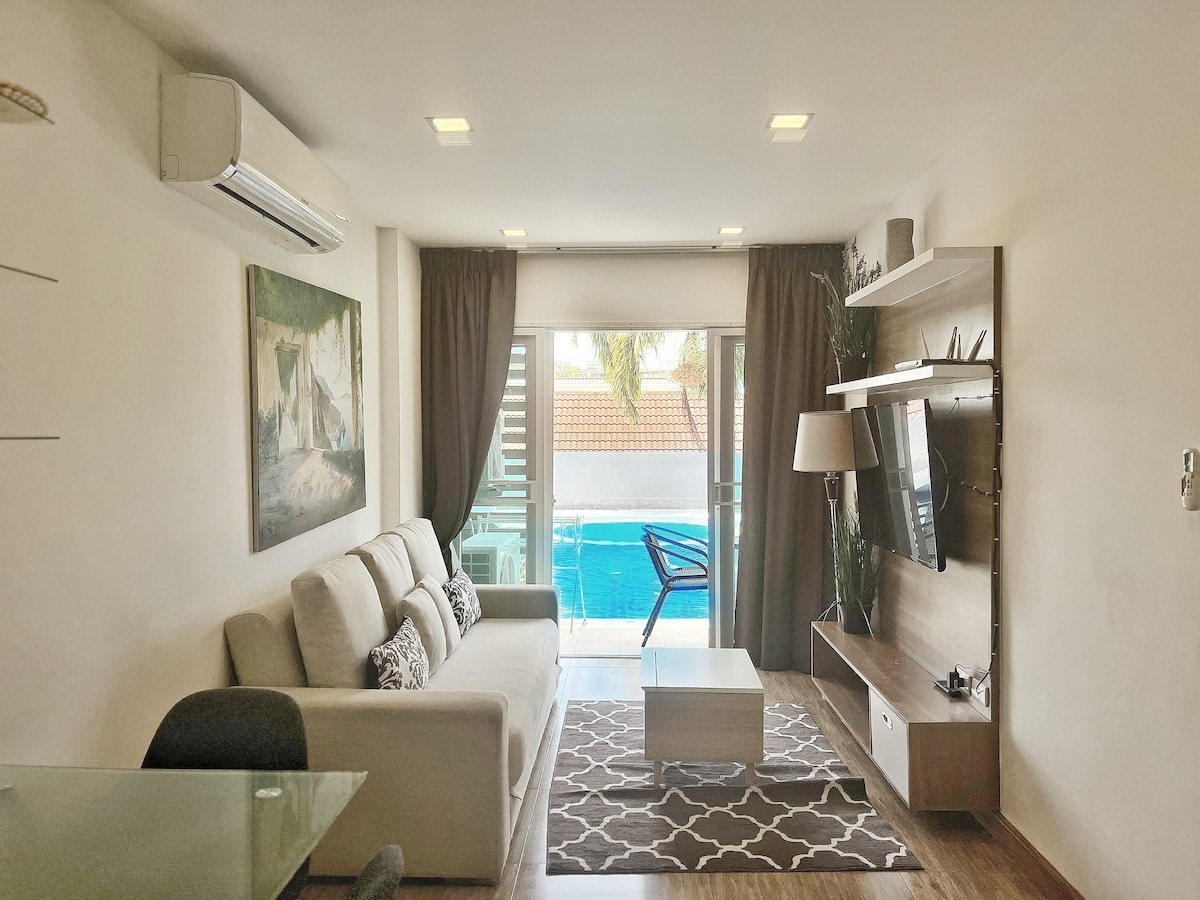 A bright living area showcases a comfortable sofa positioned near large glass doors that open to a swimming pool. A coffee table and TV unit complement the space, while neutral-toned curtains frame the windows. Decorative wall art enhances the ambiance.