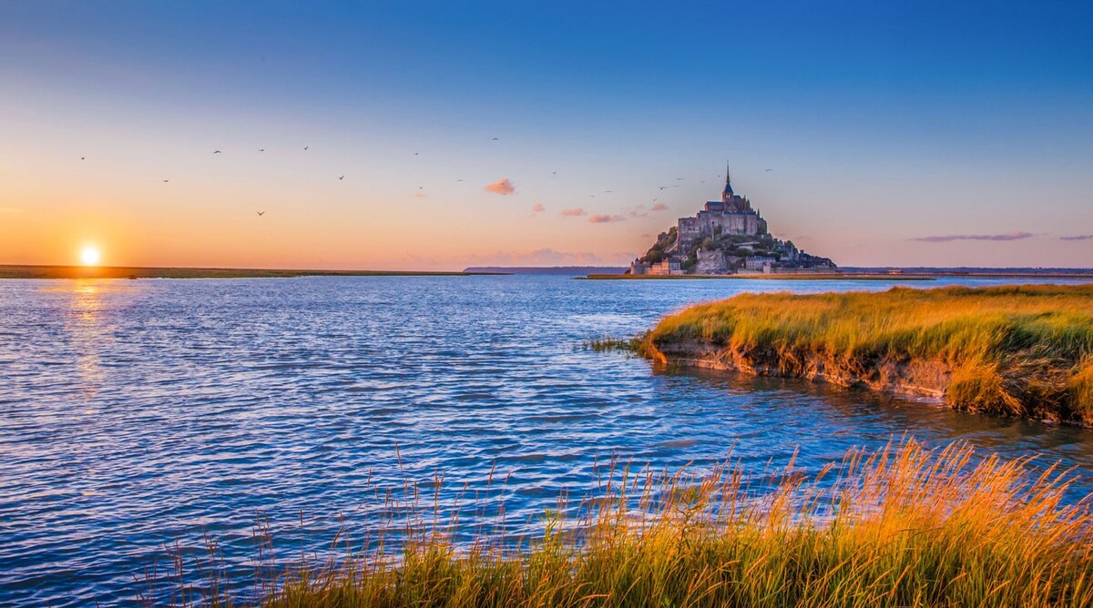 The image captures the iconic Mont Saint-Michel at sunset, surrounded by shimmering waters and gently swaying grasses. The warm hues of the sunset blend with the calm waters, creating a serene atmosphere that highlights the structure's silhouette against the colorful sky.