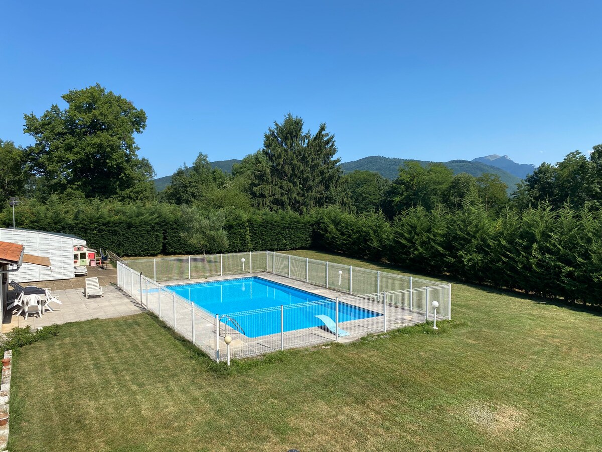 A well-maintained outdoor swimming pool is enclosed by a white fence, surrounded by a manicured lawn. Lush trees create a natural border, offering a serene backdrop of mountains under a clear blue sky. Lounge chairs are positioned nearby for relaxation.