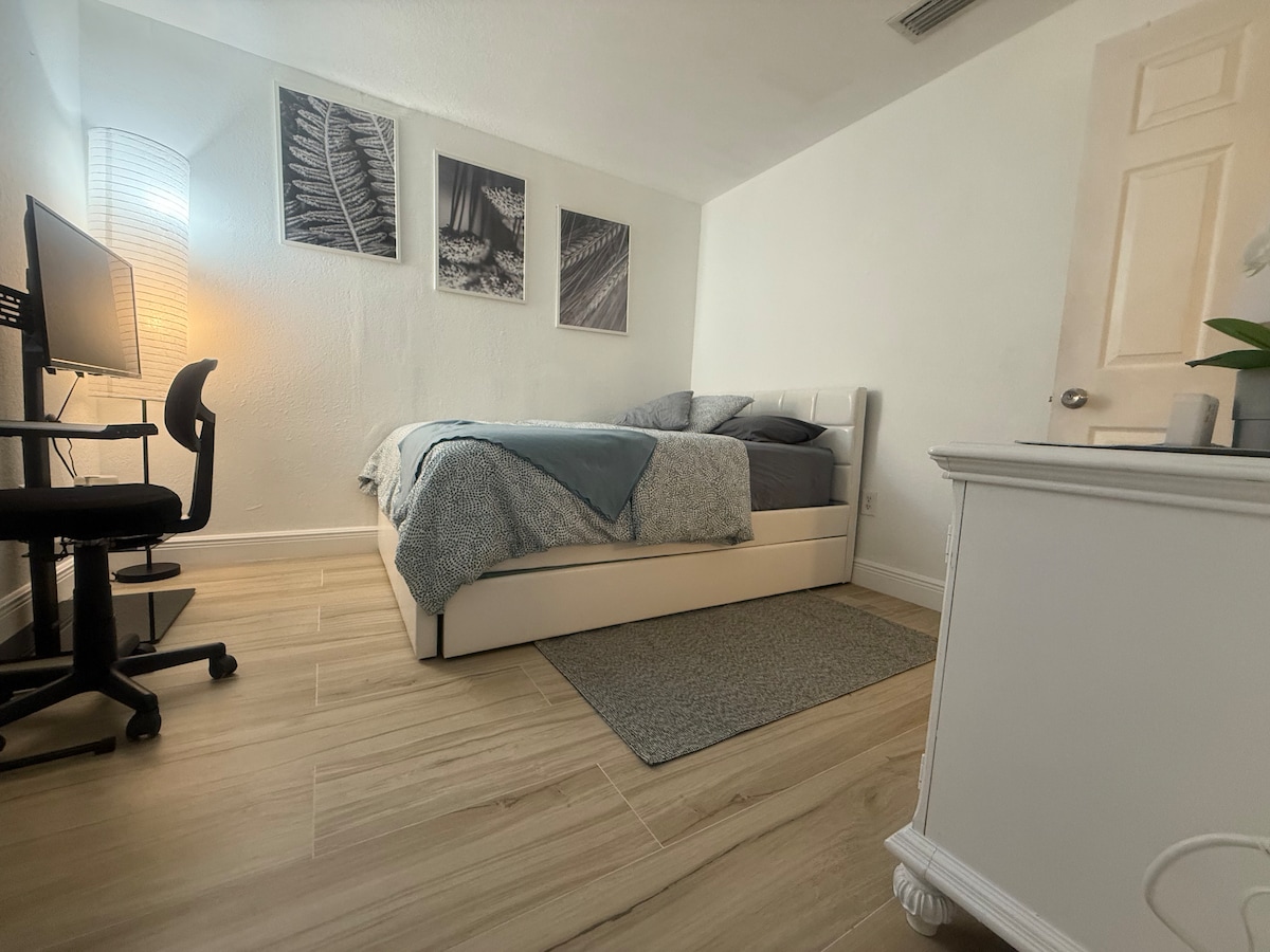 A spacious bedroom is depicted, featuring a plush bed covered with a quilted blanket and coordinating throw. Soft light emanates from a nearby lamp, illuminating the room's neutral tones. A white desk with a computer and a chair is situated adjacent to the bed, enhancing the functional layout.