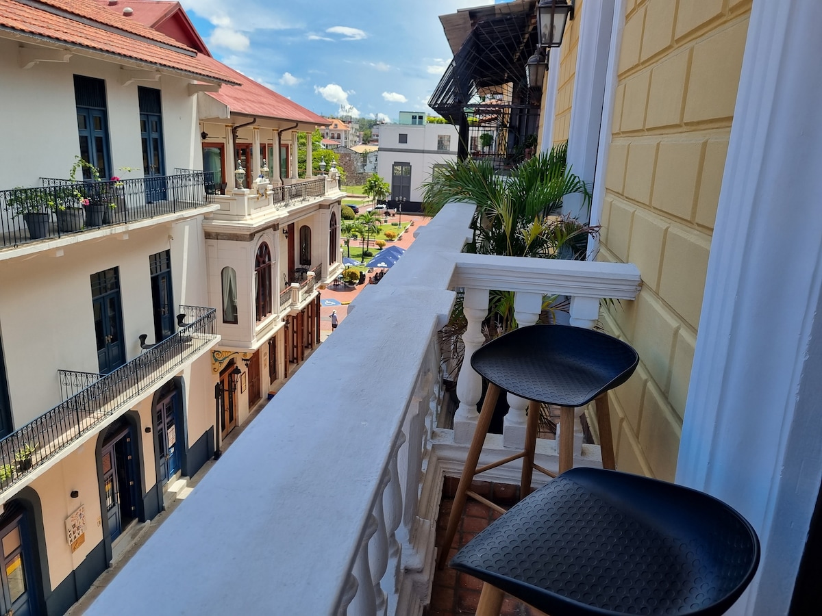A private balcony offers views of the surrounding plaza, featuring charming colonial buildings. Two black stools sit at the edge, framed by light curtains and a potted plant. Sunlight brightens the area, creating an inviting outdoor space.