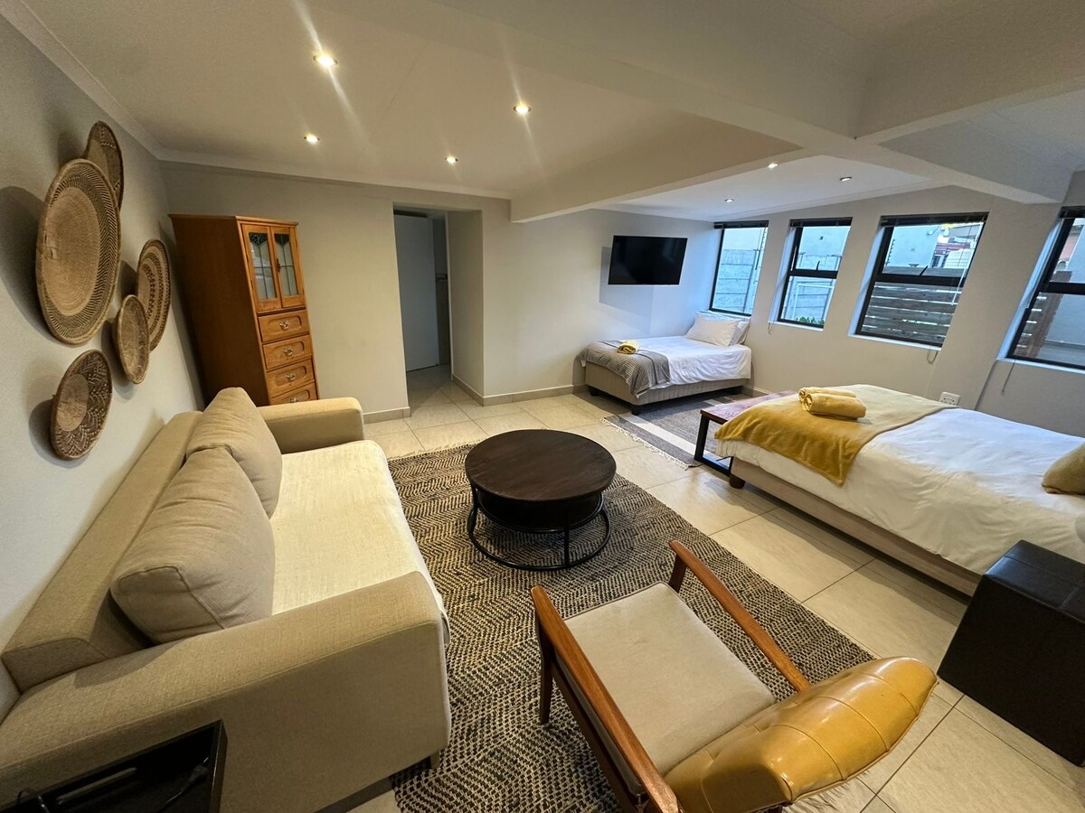 A spacious studio apartment features a lounge area with a beige sofa and a wooden armchair. Two beds, one queen size and one single, are arranged along one wall. Natural light fills the room through large windows, with decorative woven wall art enhancing the space.