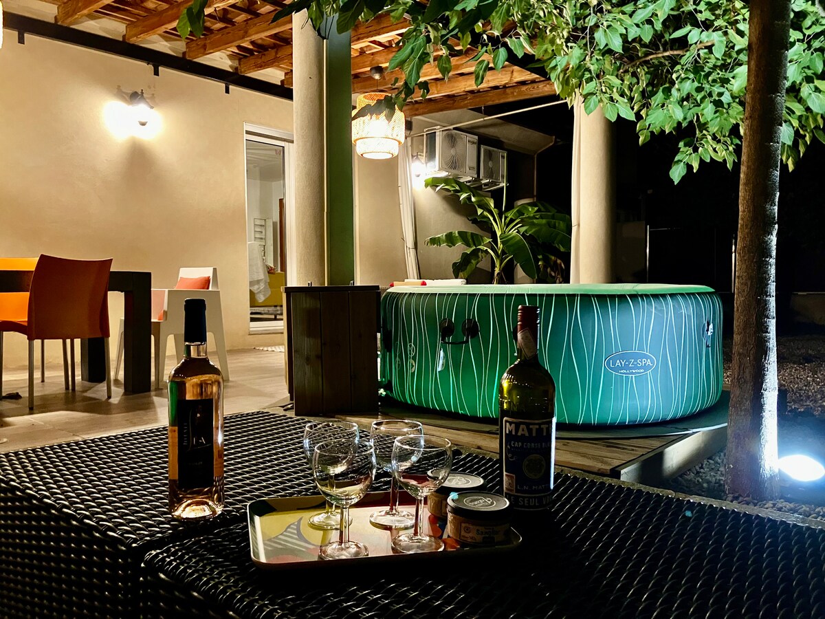 An outdoor seating area features a low table with glasses and bottles, set under a softly lit canopy. A spa jacuzzi, surrounded by foliage, is visible in the background. Vibrant chairs and a dining table provide additional seating options, creating a welcoming environment.