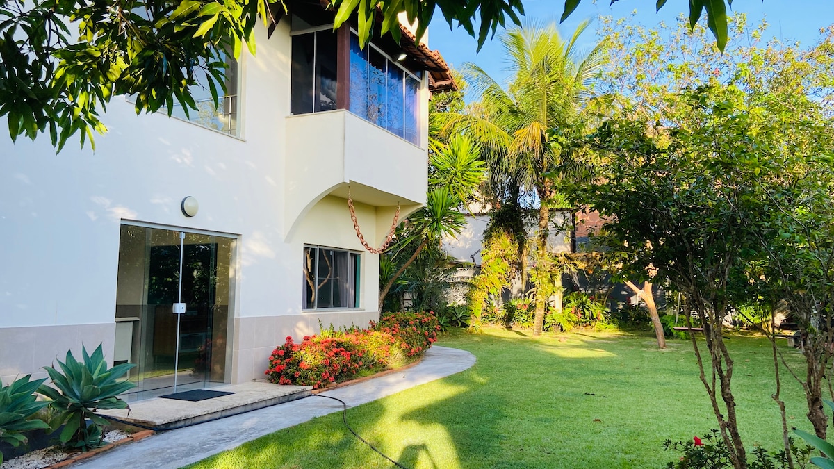 The image showcases the exterior of a duplex-style building surrounded by a well-maintained garden. Lush greenery and colorful flowers are visible, along with palm trees providing shade. A pathway leads to the entrance, which features a large glass door enhancing the warm and welcoming environment.