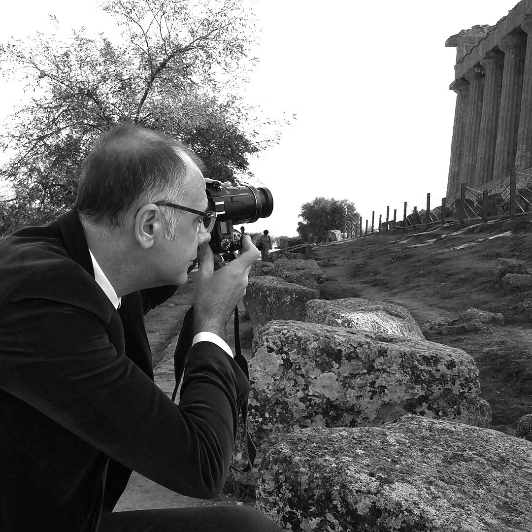 A person is seen capturing a photograph with a camera, surrounded by foliage and uneven stones. In the background, ancient structures are discernible, suggesting a historical site. The environment appears serene, with natural elements contributing to a tranquil atmosphere.
