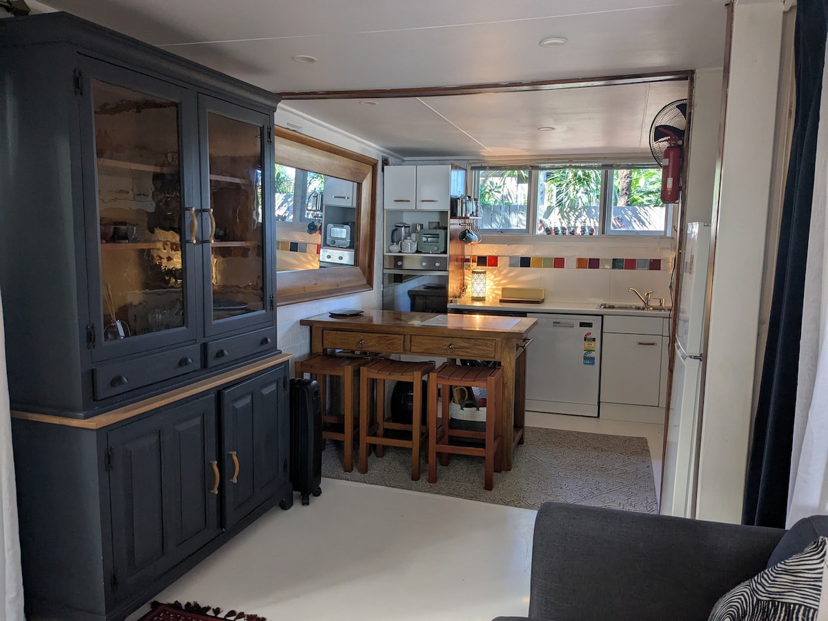 A functional kitchen area features wooden dining furniture, set against a wall adorned with colorful tiles. Modern appliances are neatly arranged, and large windows allow natural light to illuminate the space. A cozy seating area is visible, contributing to the inviting atmosphere.