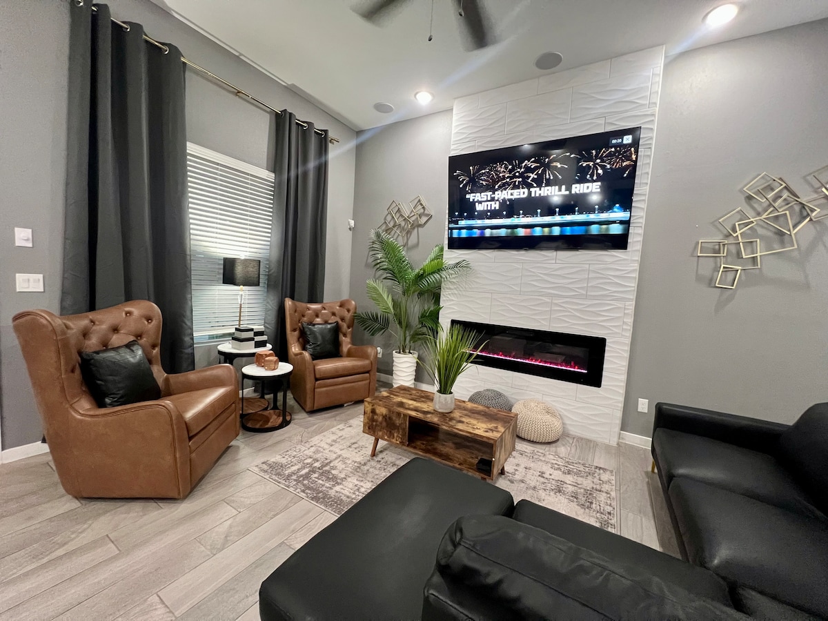 A contemporary living area is featured, displaying brown leather armchairs paired with a sleek black sectional sofa. A stone fireplace adds warmth, while a mounted TV presents an engaging display. Soft lighting from a table lamp and natural light from a nearby window enhance the inviting space.