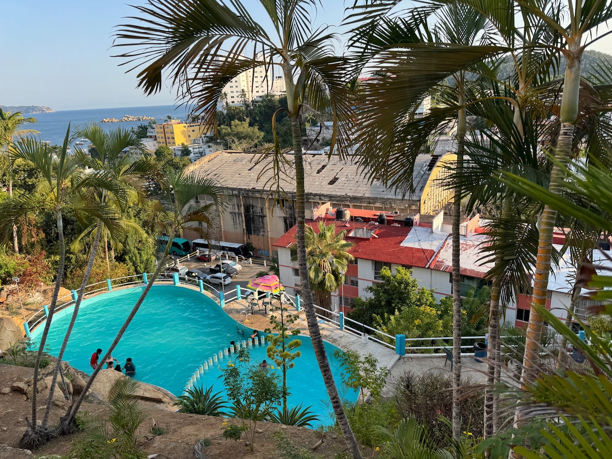 A serene pool area is surrounded by palm trees and lush greenery, offering a view of the nearby coastline. The pool features a gentle curve and is accompanied by landscaped gardens. Buildings are visible in the background, highlighting the inviting atmosphere of the space.