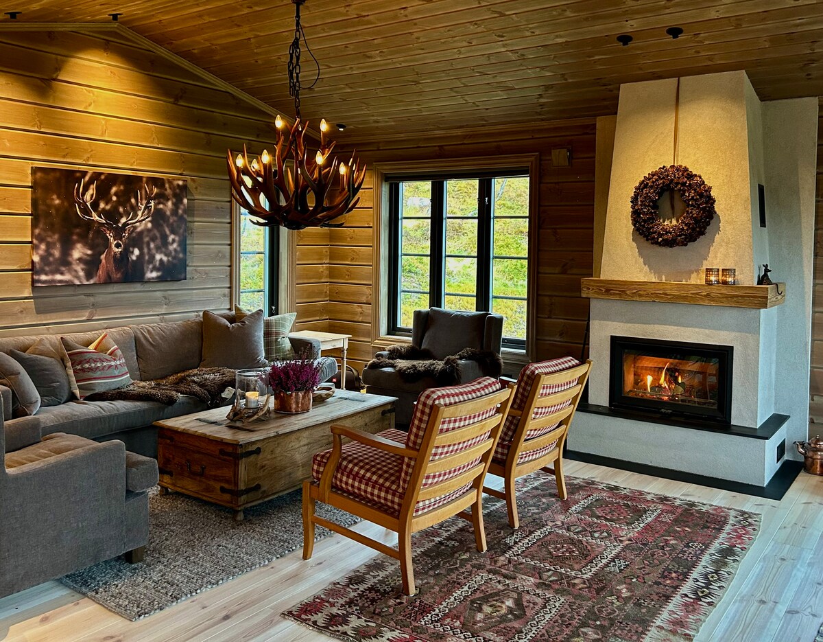 A cozy living area features a large sofa, a rustic coffee table, and two wooden chairs with patterned upholstery. A warm fireplace is positioned against a light-colored wall, while large windows allow natural light to fill the space, complemented by a decorative chandelier.