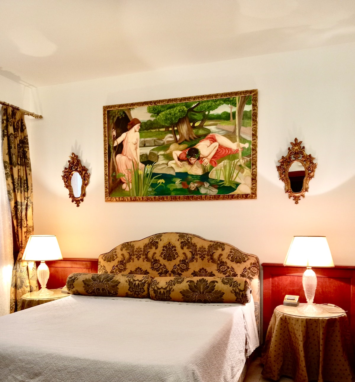 A serene sleeping area is highlighted by a spacious bed adorned with a soft white coverlet. Decorative wall mirrors and nightstands with lamps enhance the inviting space. A large artwork depicting a tranquil scene is mounted above the bed, echoing classic Venetian style.