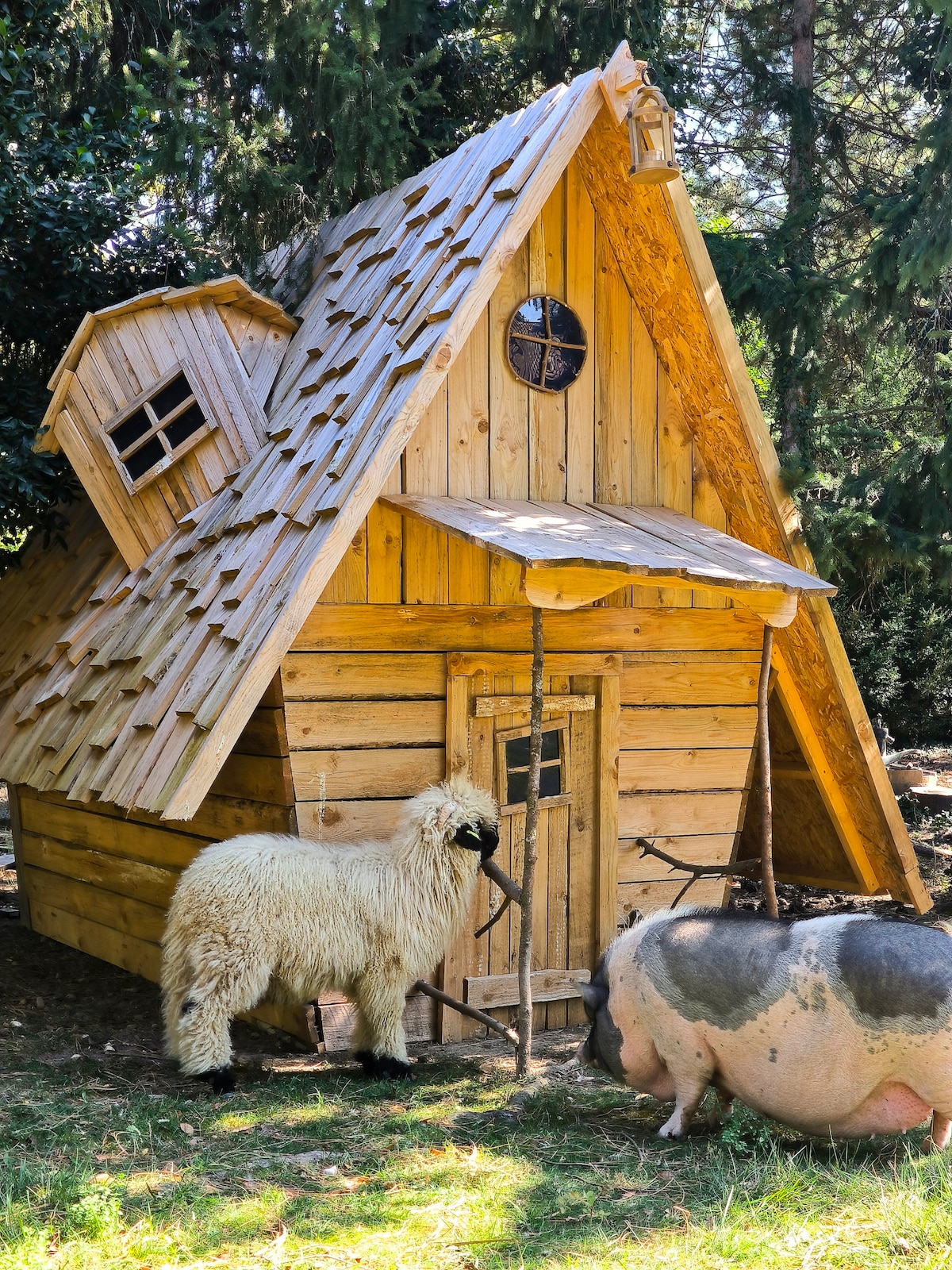 A rustic wooden structure resembling a small cottage is presented with a sloped roof and a round window. Nearby, a sheep stands next to a small pig, both situated on a grassy area surrounded by trees.
