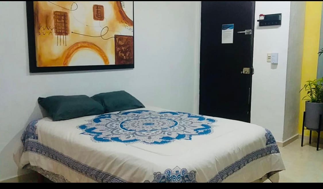 A spacious bed is centered in the room, adorned with a decorative blanket featuring a blue mandala design. Two dark green pillows rest at the head of the bed. An abstract artwork in warm tones hangs above the bed, complementing the room's neutral walls.