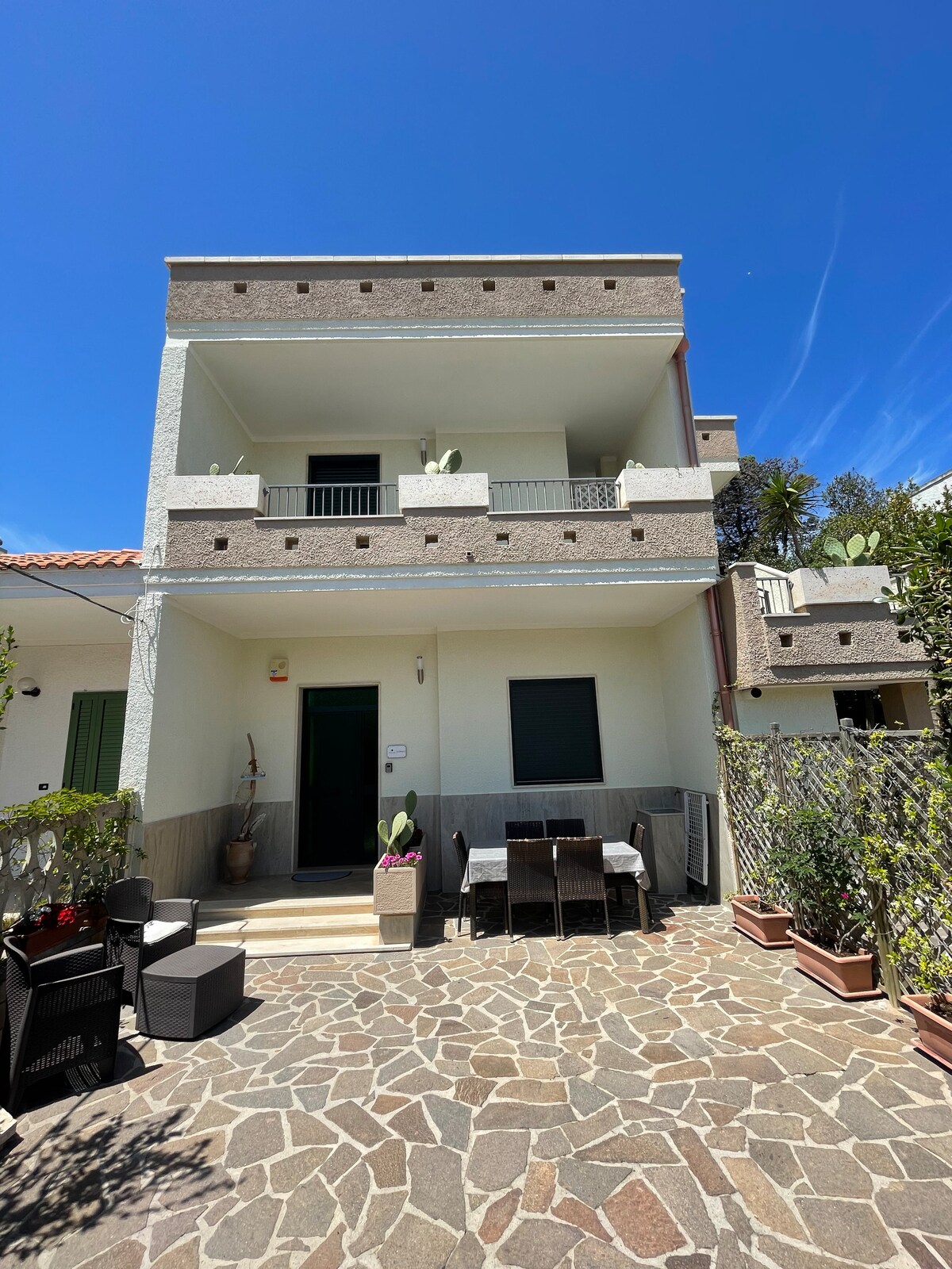 The exterior view of the two-story apartment features a light-colored facade complemented by green accents. A small outdoor seating area is present, with a table and chairs. Lush potted plants and a stone pathway frame the entrance, leading to spacious balconies on the upper level.