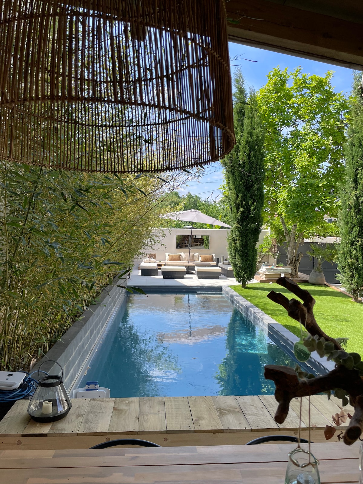 A private pool reflects the clear blue sky, surrounded by lush greenery and tall cypress trees. A shaded lounging area is visible at the far end of the pool, furnished with comfortable seating. Sunlight dapples the well-maintained garden, enhancing the serene outdoor space.