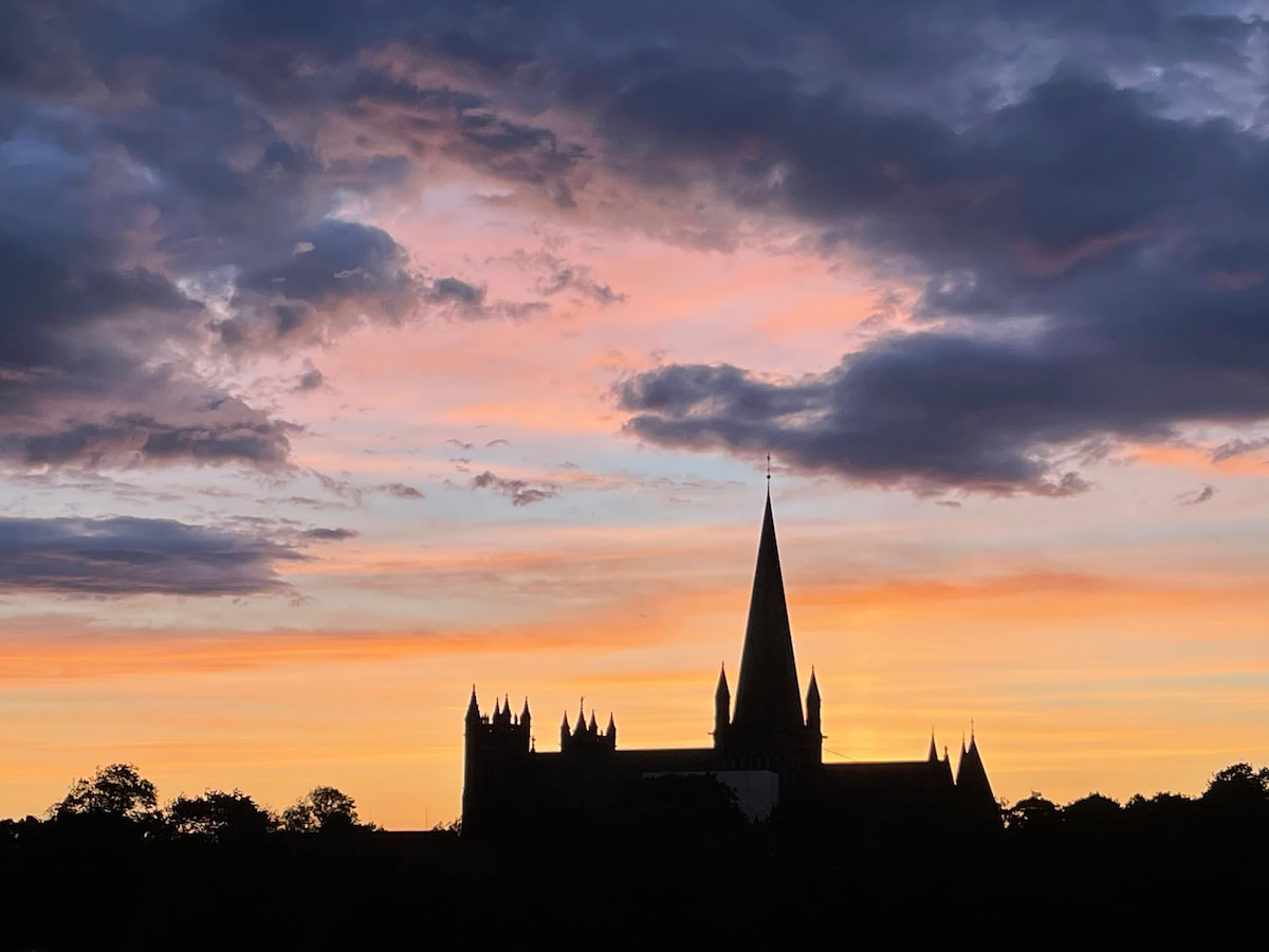 A silhouette of a historic cathedral is depicted against a colorful sunset sky, with soft hues of orange, pink, and purple. The sharp peak of the cathedral's steeple stands prominently among a backdrop of clouds, creating a serene evening scene.