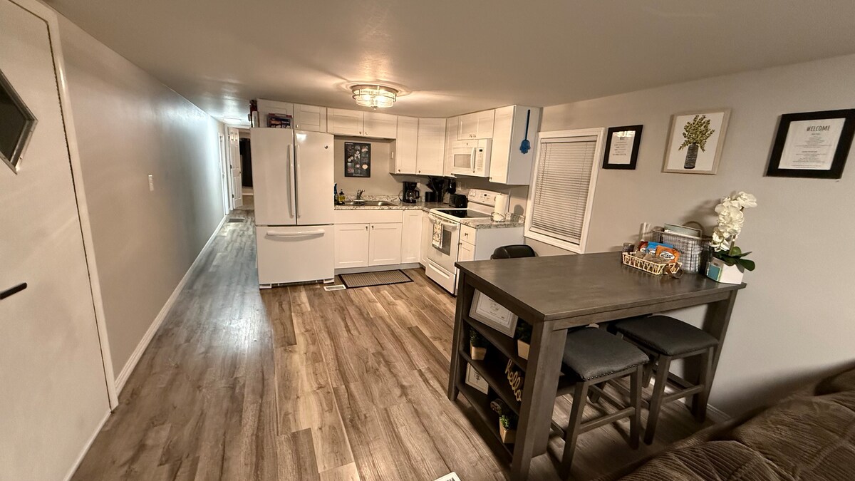 A modern kitchen space is visible, featuring white cabinetry and stainless steel appliances. A wooden dining table with dark chairs is set in the foreground. Natural light enters through a window, illuminating the light-colored flooring that stretches toward a hallway with additional doors.
