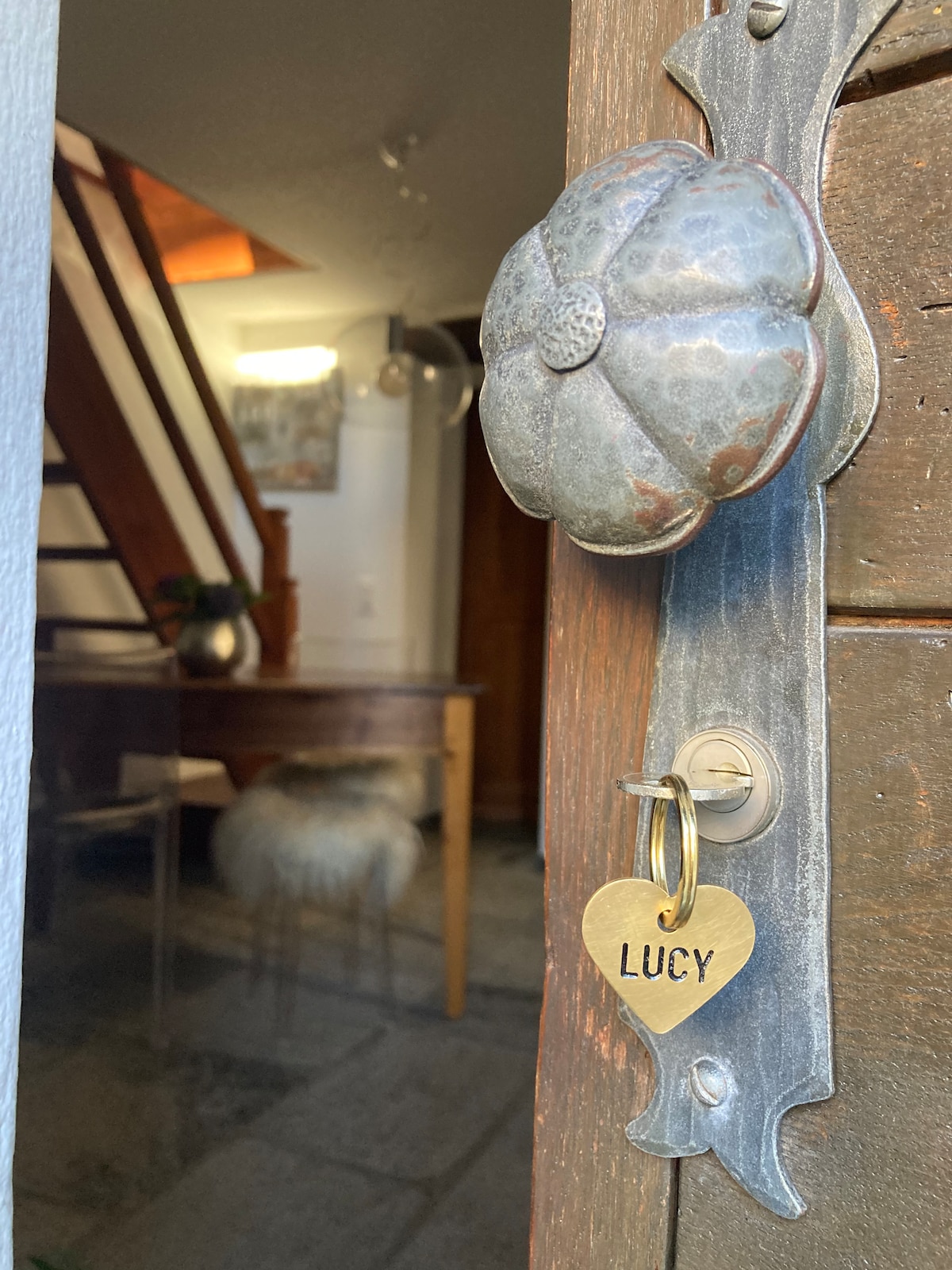 A close-up view of the rustic wooden door features a decorative handle and a heart-shaped keychain labeled 'LUCY.' The doorway leads into a cozy interior space with visible stairs and a dining area in the background.
