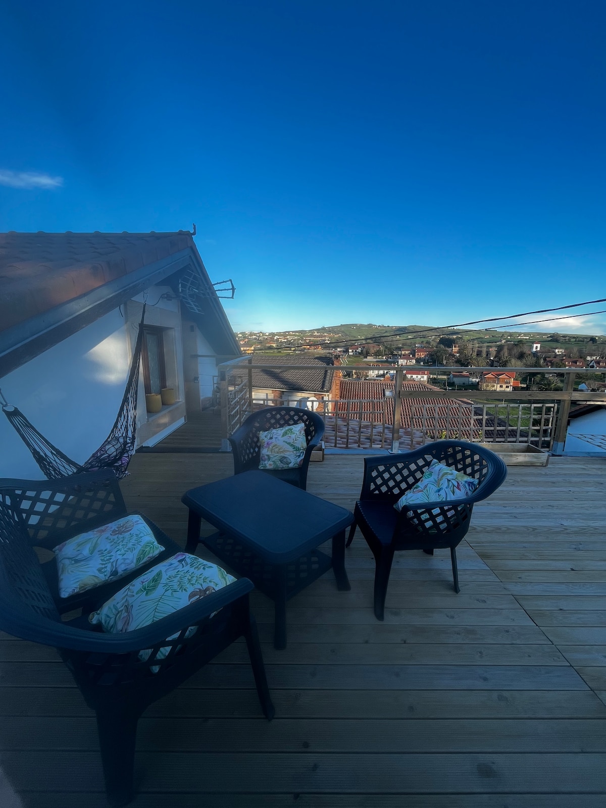 A sunlit terrace features a small table surrounded by four black chairs with patterned cushions. A hammock hangs nearby, creating a relaxing space with views of the surrounding landscape and clear blue sky.