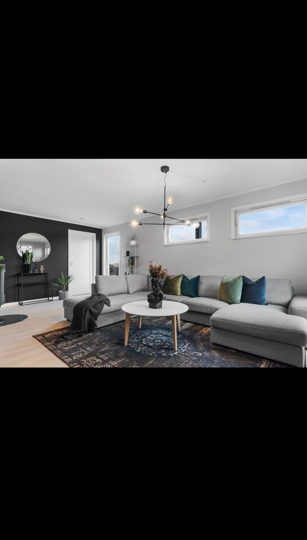 A modern living area features a light gray sectional sofa with colorful cushions. A round coffee table is placed in front, with a small plant resting on it. Natural light enters through two windows, enhancing the space's open feel. A decorative mirror and a dark accent wall add depth.